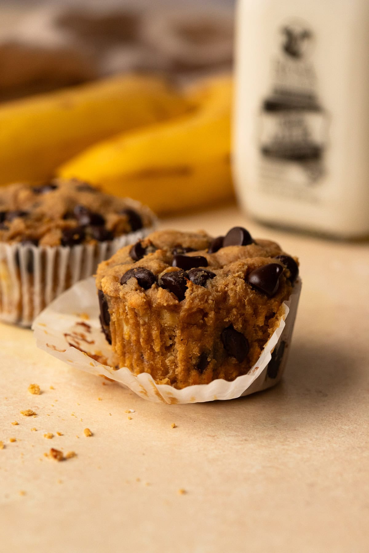 A muffin with the paper liner partially removed and bananas in the background.