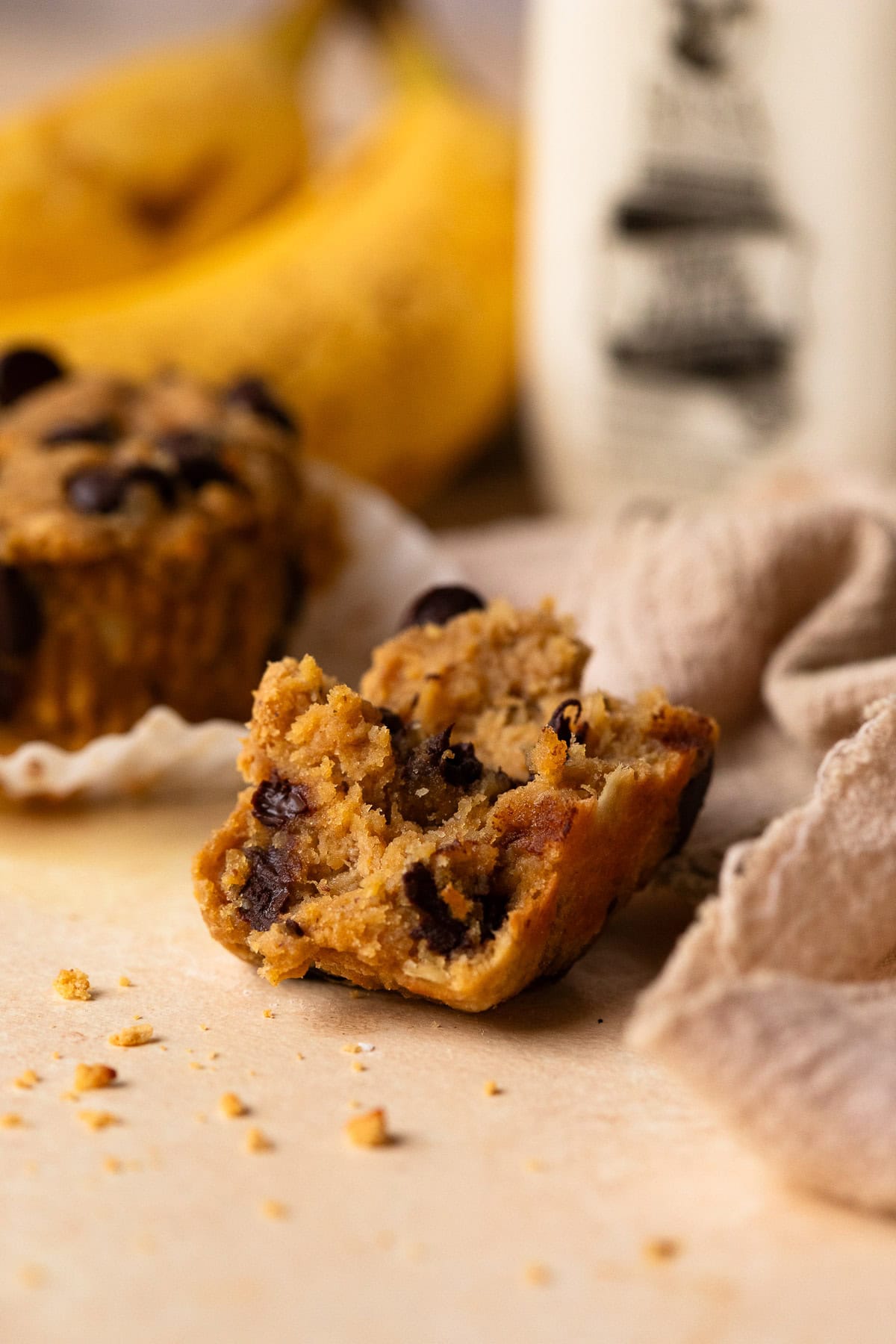 A piece of a muffin with a full muffin, bananas, and a glass milk bottle in the background.